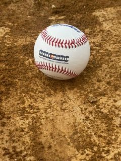 A baseball resting on a dirt infield, showing red stitching and a logo on the side.
