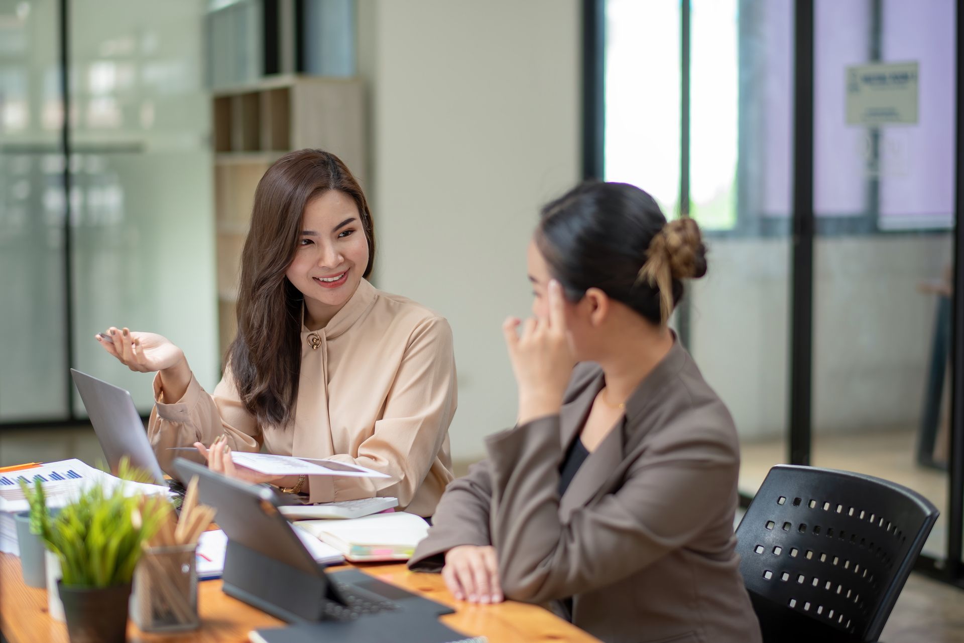 Two women are sitting at a table with laptops and talking to each other.