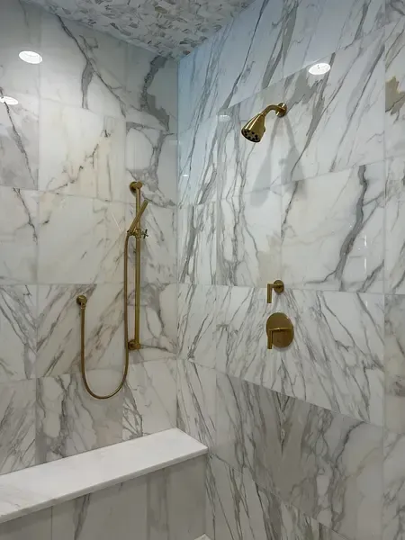 Marble-tiled shower with gold fixtures, handheld sprayer, wall shower head, and bench seat