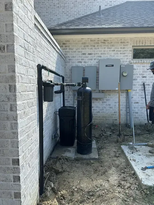 Outdoor water treatment equipment beside a brick house, including tanks, filters, and utility boxes.