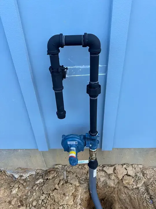 Outdoor water meter and black PVC pipes mounted on a light blue wall above a dirt ground