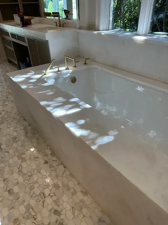 Modern bathroom with a white bathtub, gold fixtures, and a stone tile floor lit by sunlight.