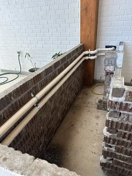Partially built brick utility trench with exposed pipes along a white wall