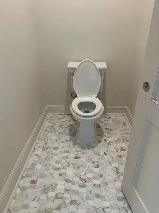 Small bathroom with an open toilet, beige walls, and patterned tile floor.