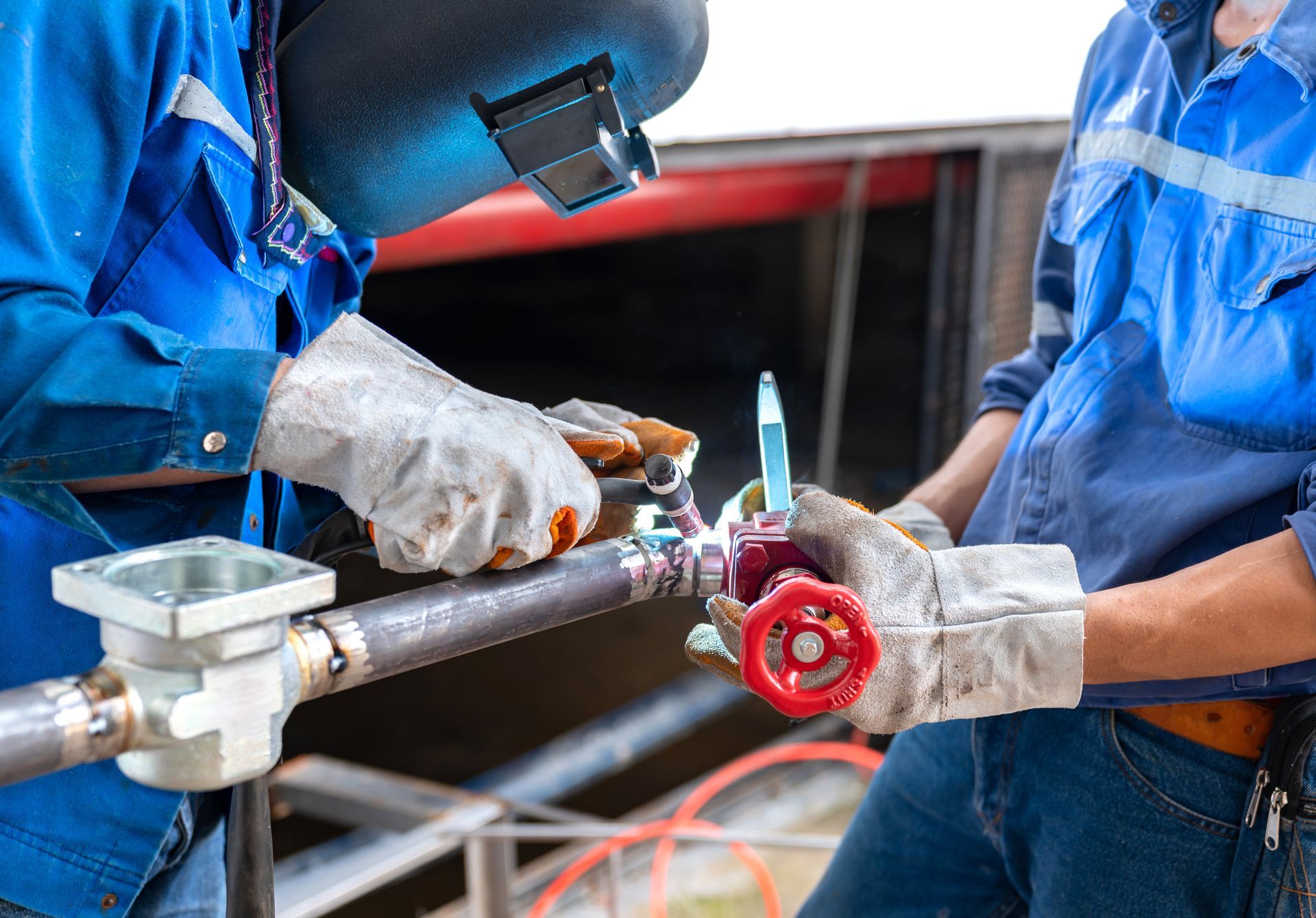 Two workers in blue coveralls inspecting a pipe valve with tools in an industrial setting