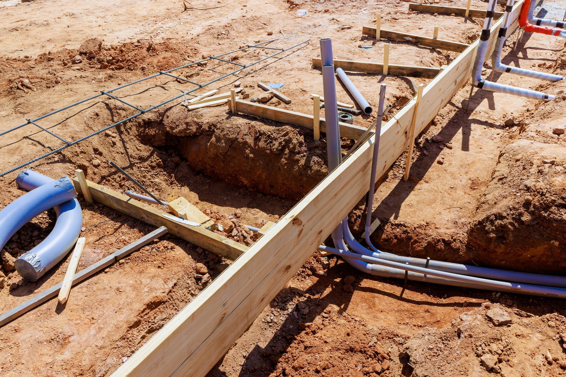 Construction site with trenches, wooden forms, and blue utility pipes in dry soil