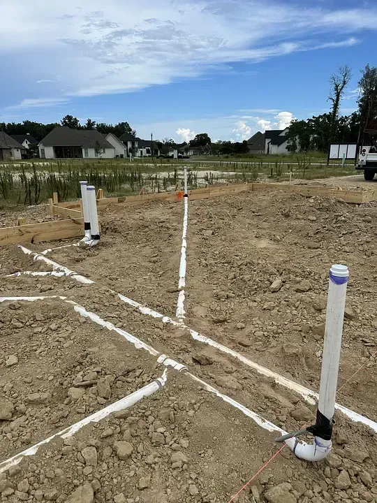 Marked dirt lot with white boundary stakes and string under a blue sky, near houses and a field