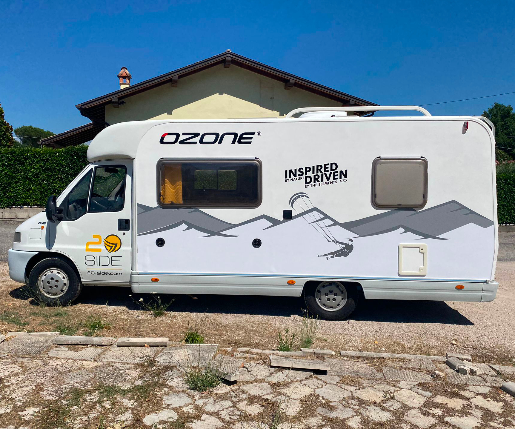 Un camper Ozone bianco con grafiche di montagna e parapendio parcheggiato su una superficie ghiaiosa di fronte a una casa.