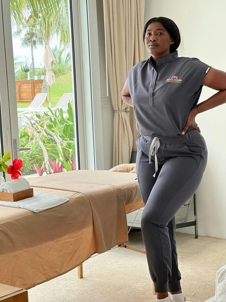 Person in gray athleisure posing beside a massage table in a bright room with large windows.