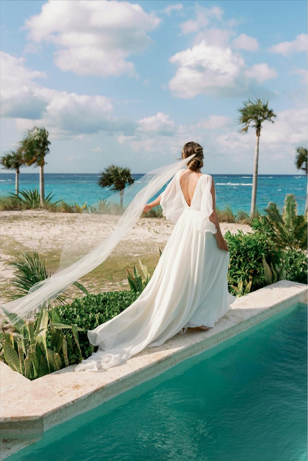 Bride in a flowing white gown and veil standing by a pool overlooking a tropical beach and ocean