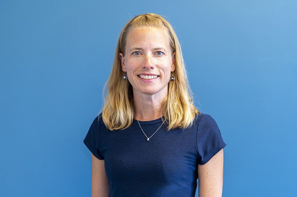 Woman with blonde hair wearing a dark blue shirt smiles at the camera, standing in front of a blue wall.