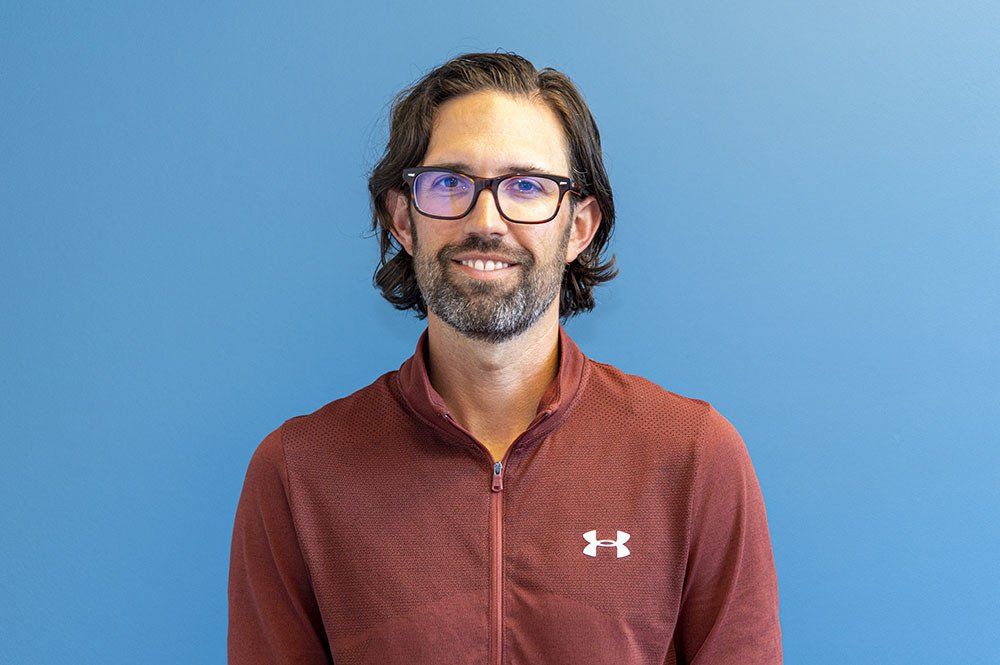 Man with glasses and beard, wearing a red zipped top, smiling against a blue backdrop.