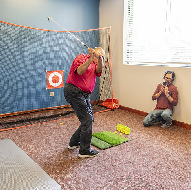Man swinging a golf club indoors toward a net; another person kneels taking a photo.