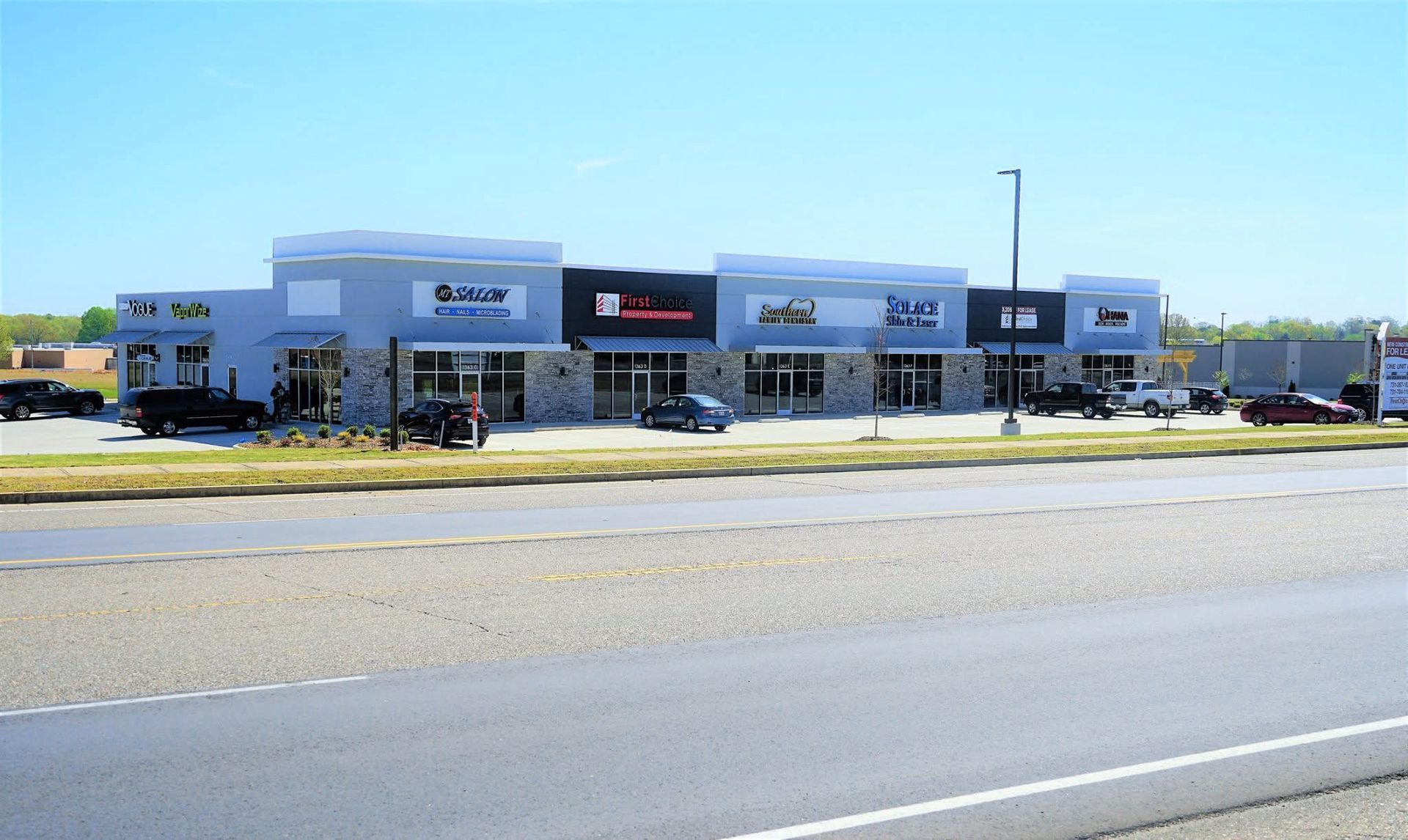 A strip mall with multiple businesses, light blue and stone facade, parked vehicles, and a clear sky.