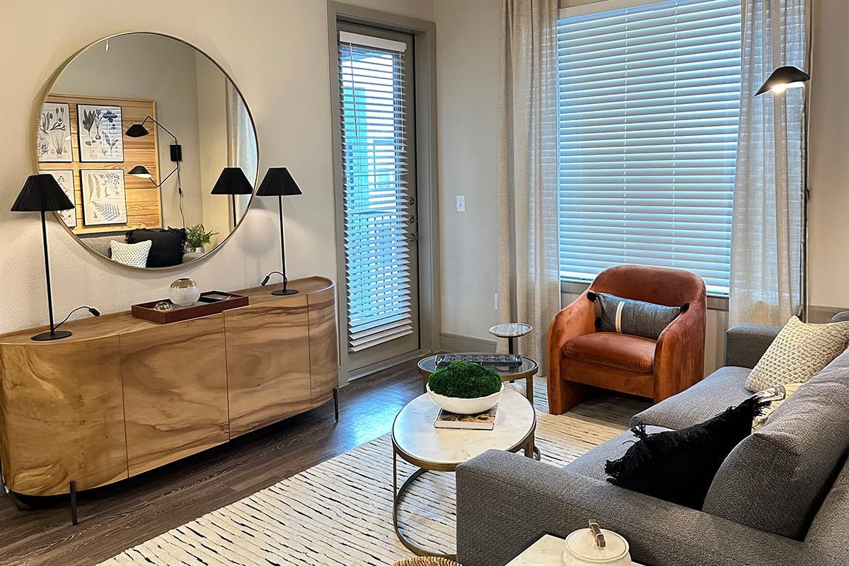Living room in a modern apartment with a curved wood sideboard, round wall mirror, sofa, and orange chair.