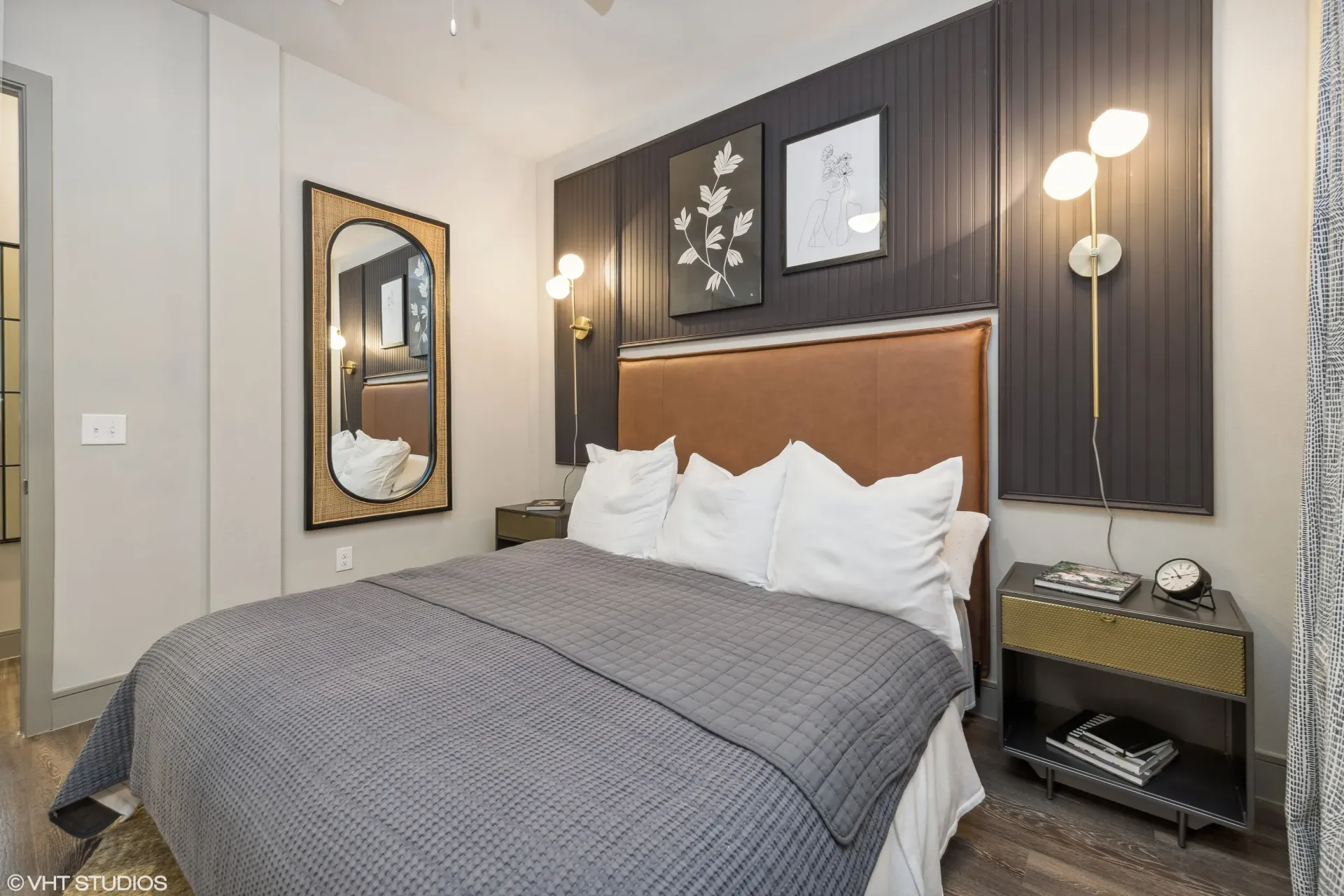 Bedroom inside an apartment with a brown upholstered headboard, gray bedding, nightstands, and framed wall art.