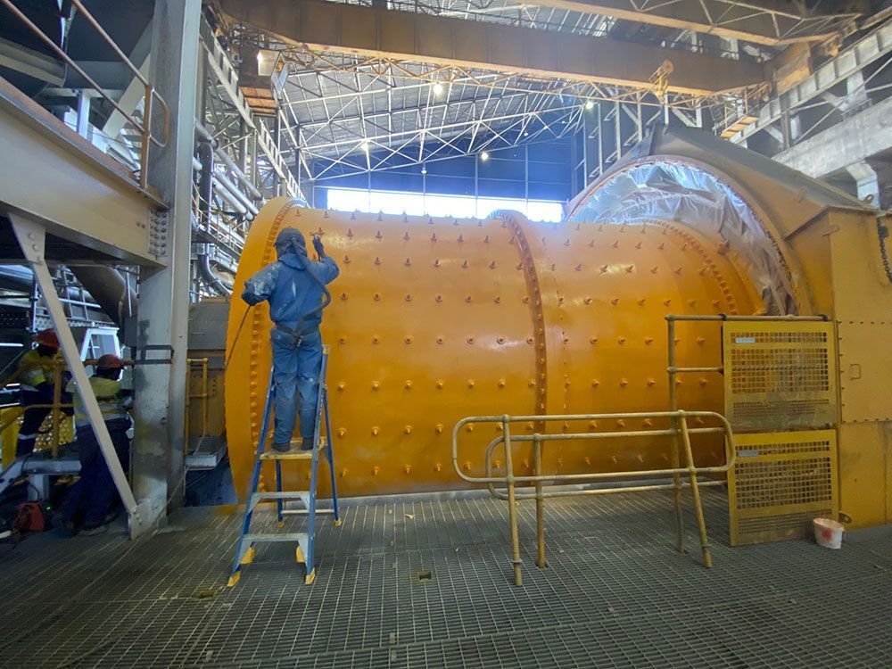A Man Is Standing On A Ladder In Front Of A Large Yellow Pipe Applying Protective Coating — Procoat QLD in Ryan, QLD