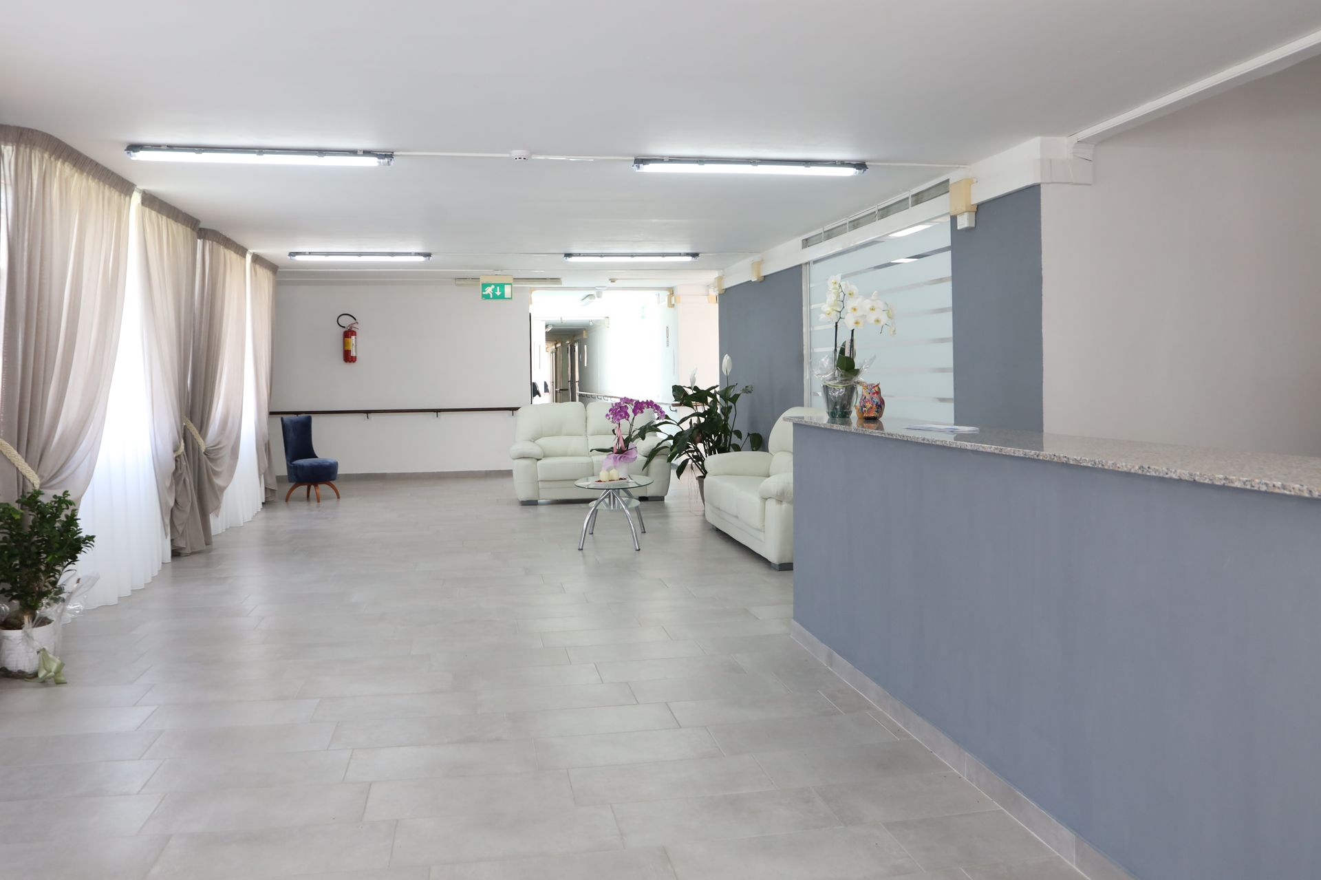 Ampia hall con pareti grigio chiaro, divani bianchi, bancone della reception e piante in vaso.