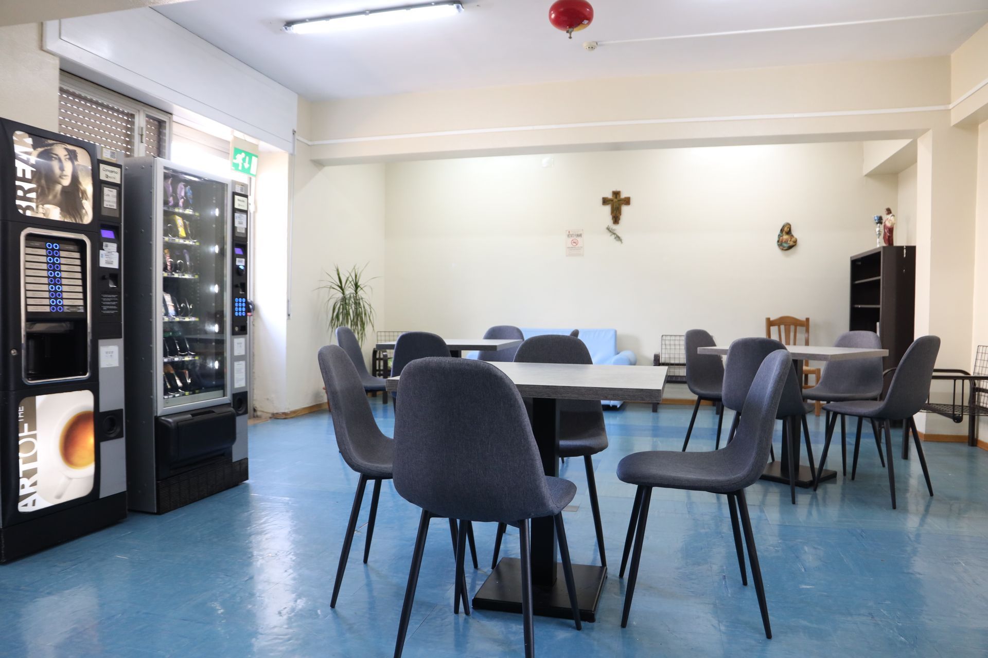 Caffetteria con pavimento blu, tavoli, sedie, distributori automatici e alcune decorazioni alle pareti.