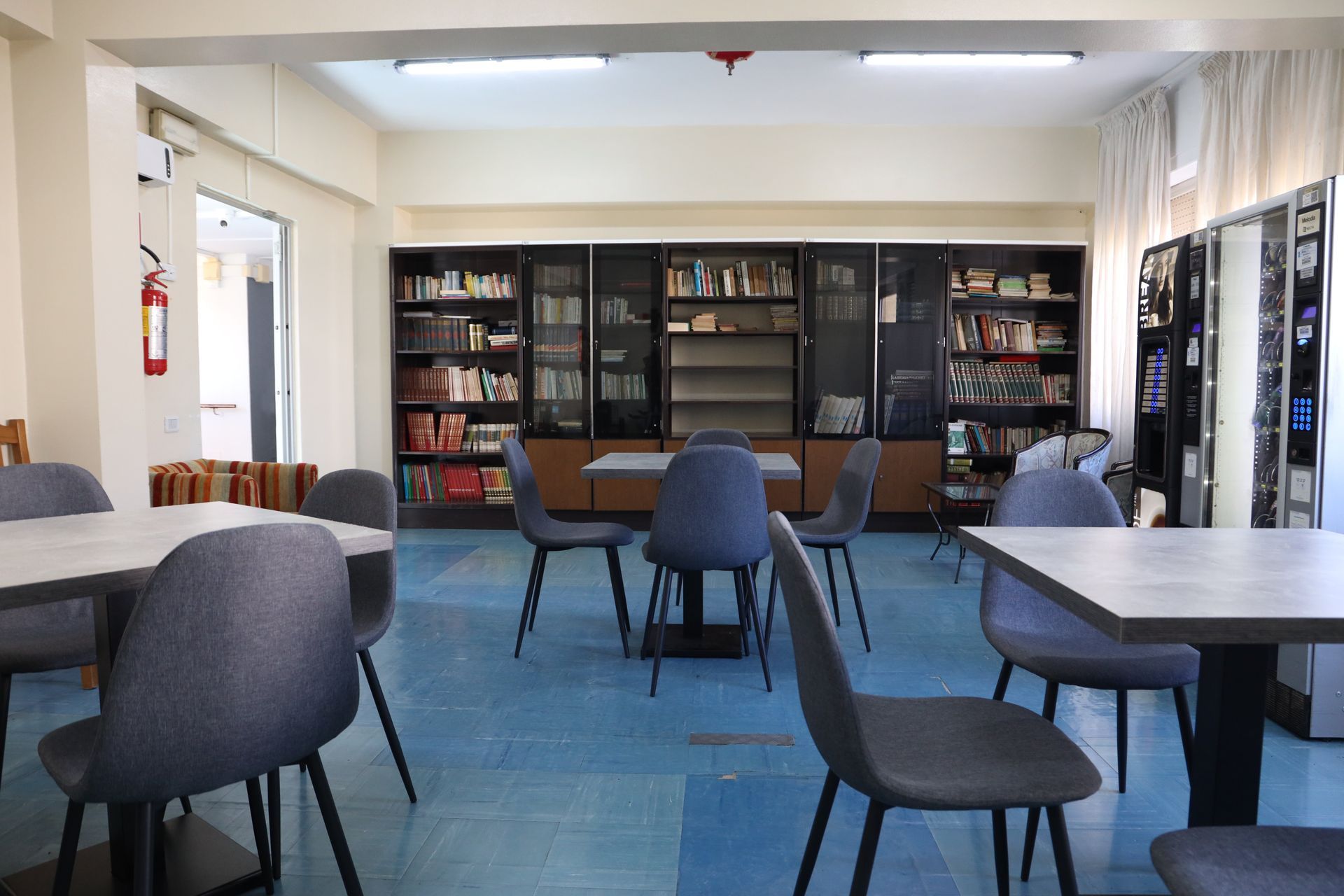 Interno di una biblioteca con tavoli, sedie e librerie. Pavimento blu, pareti chiare.