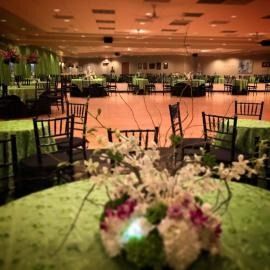Decorated banquet hall with green tablecloths, black chairs, and floral centerpieces on a polished floor