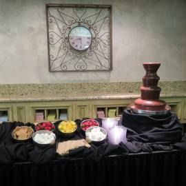 Table with bowls of colorful treats beside a stacked chocolate fountain and decorative wall clock