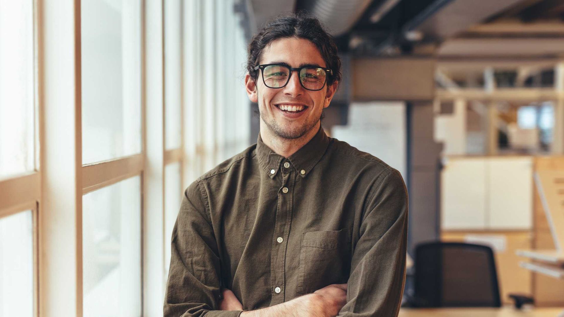 A man wearing glasses is standing in front of a window with his arms crossed and smiling.