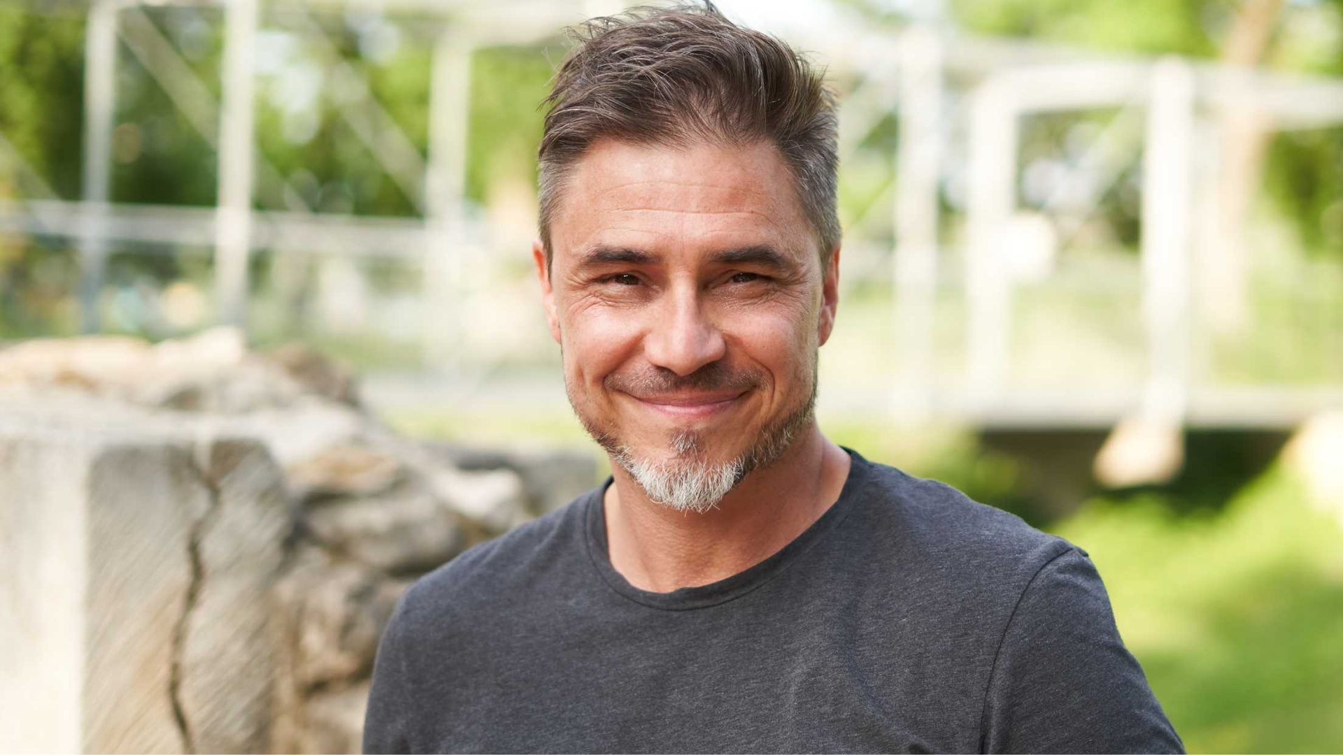 A man with a beard is smiling for the camera while standing in front of a bridge.