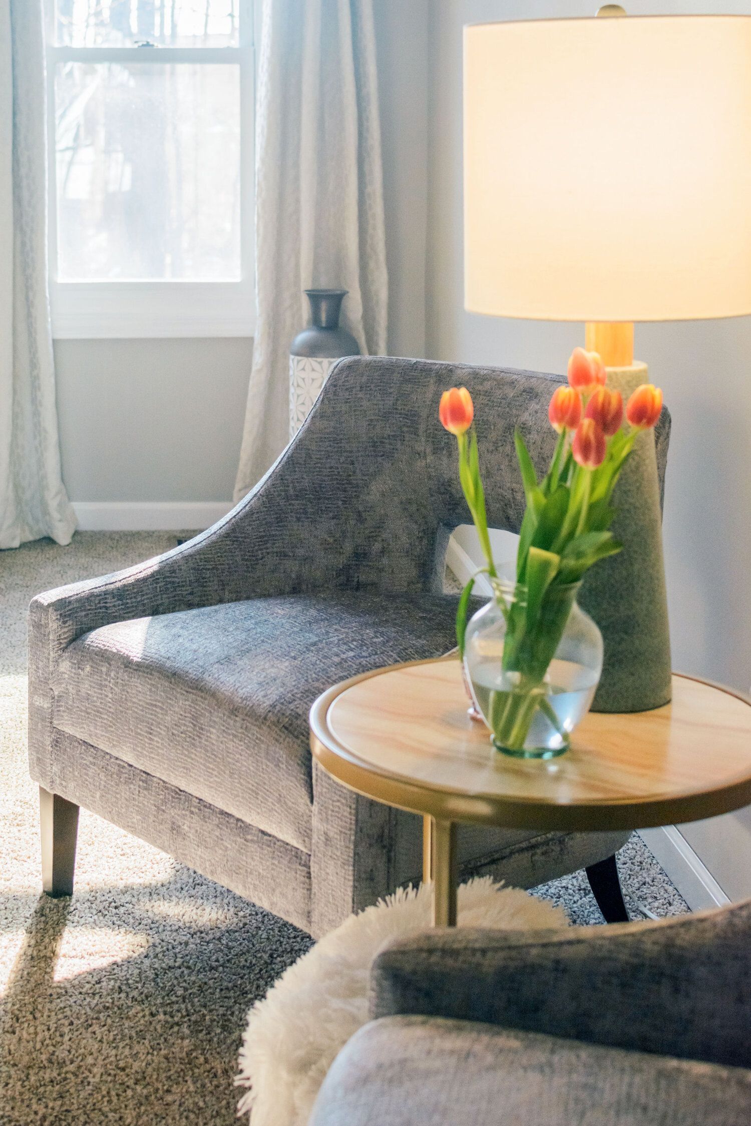 A living room with a chair , table , lamp and vase of flowers.