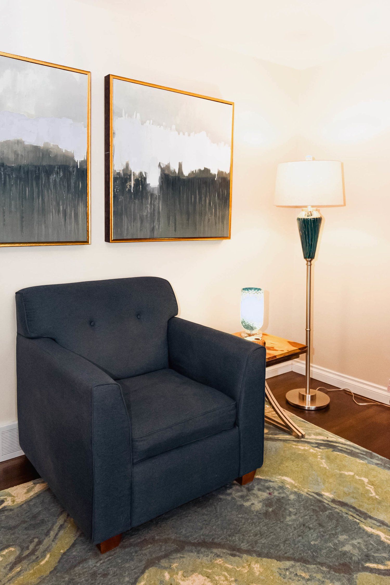 A living room with a chair , lamp , table and paintings on the wall.