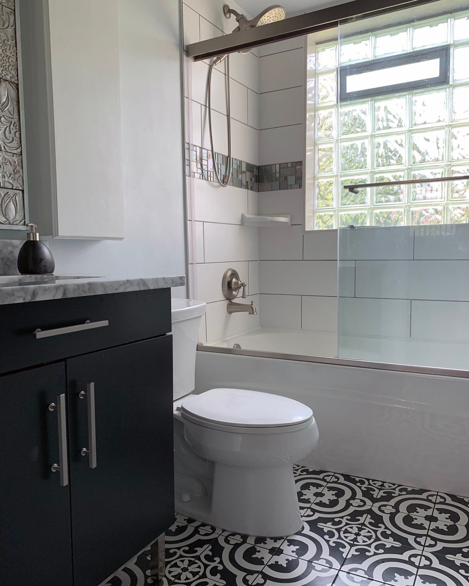 A small bathroom featuring a black vanity, a white toilet, and a glass-block window above a white tiled shower-tub.