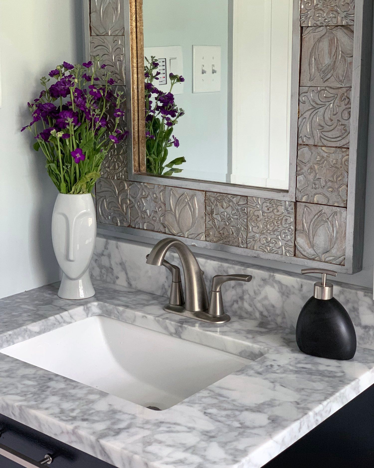 A bathroom vanity with a marble countertop, brushed metal faucet, a decorative tiled mirror, and a purple floral vase.