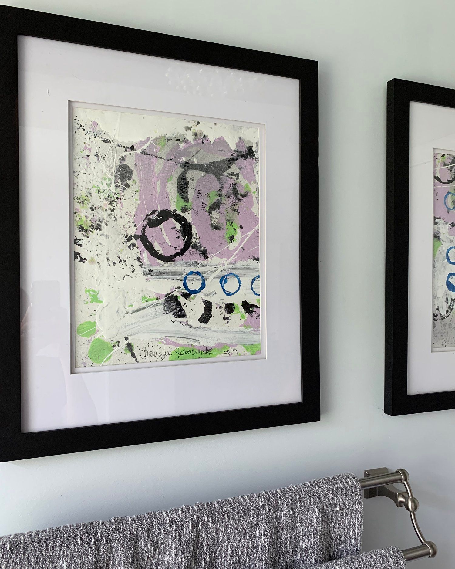 Two framed abstract paintings with purple, green, and black splatters hanging above a towel bar on a light wall.
