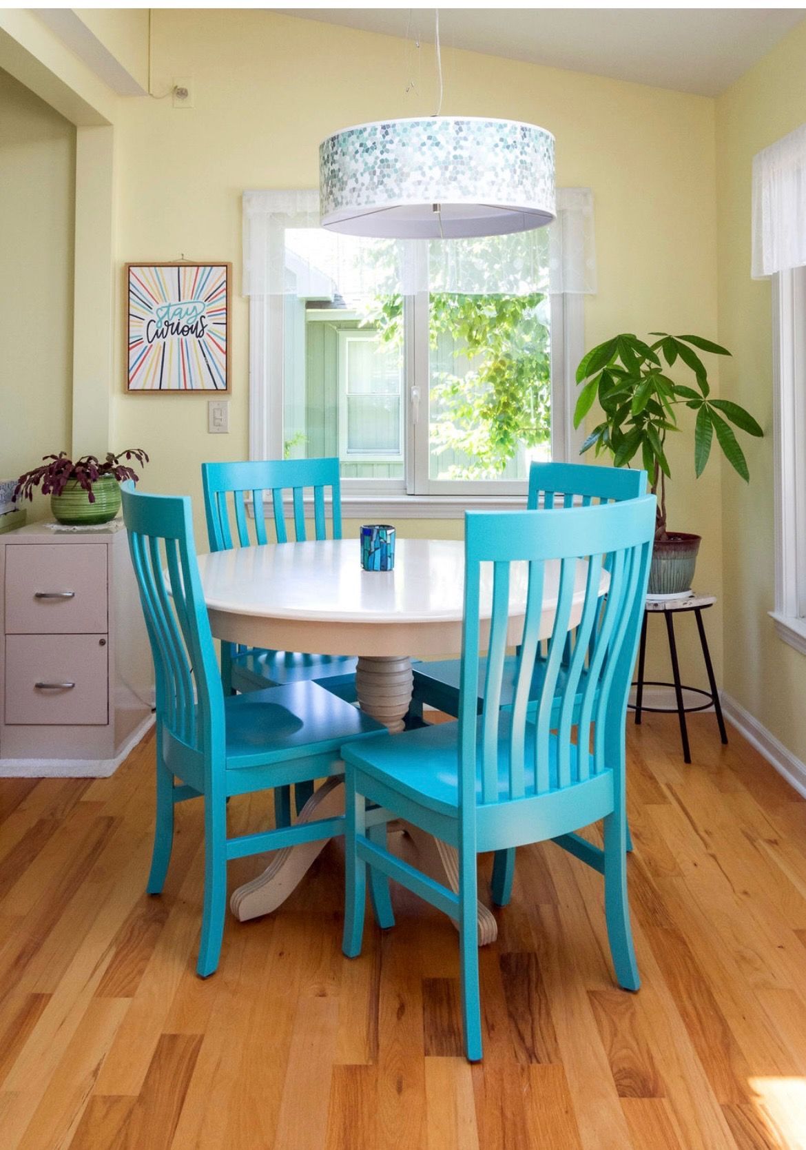 A small dining area with a round white table, four bright teal chairs, a patterned pendant light, and a potted plant.