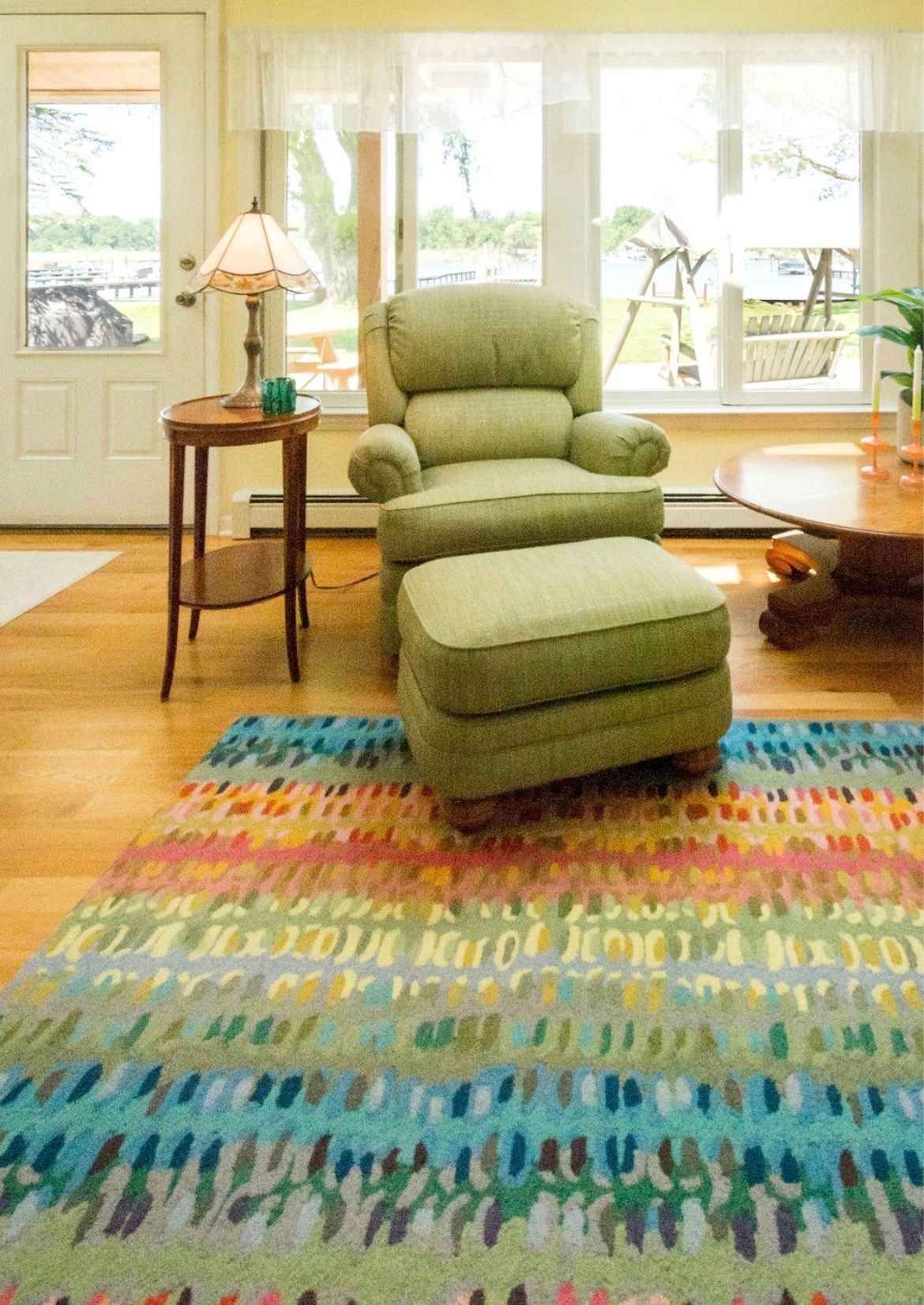 A light green armchair and matching ottoman sit on a colorful, patterned rug in a sunlit room near a large window.