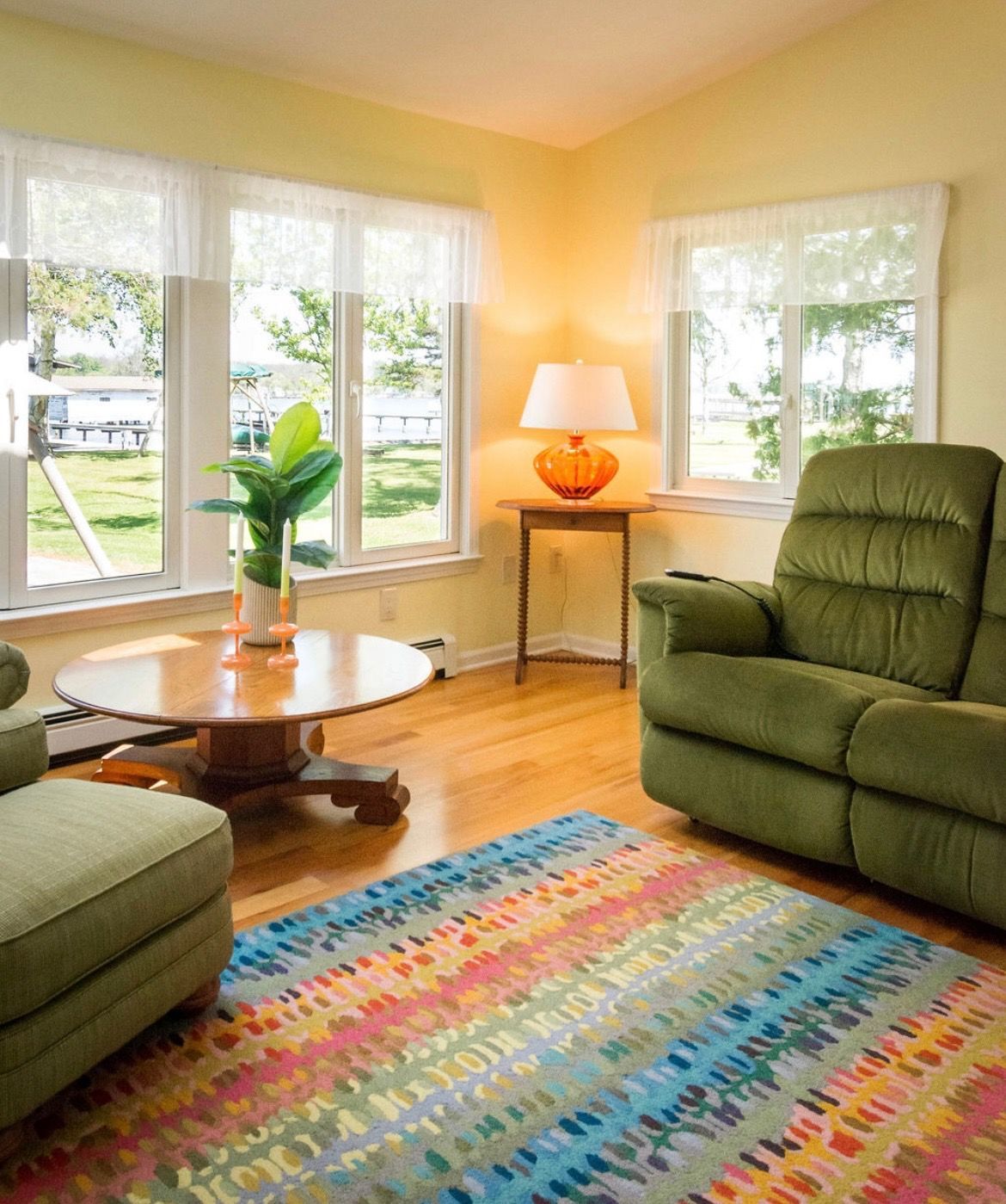 A sunlit living room featuring two green armchairs, a round wooden coffee table, a colorful rug, and a lamp on a side table.
