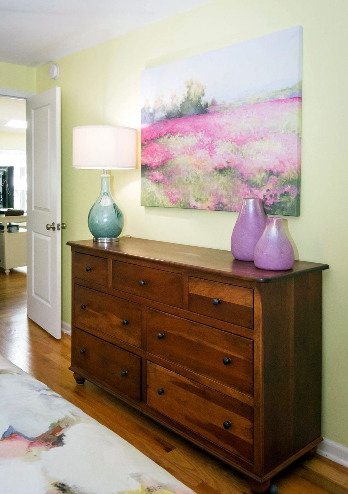 A wooden dresser with a blue lamp and two purple vases beneath a painting of a pink landscape on a pale green wall.
