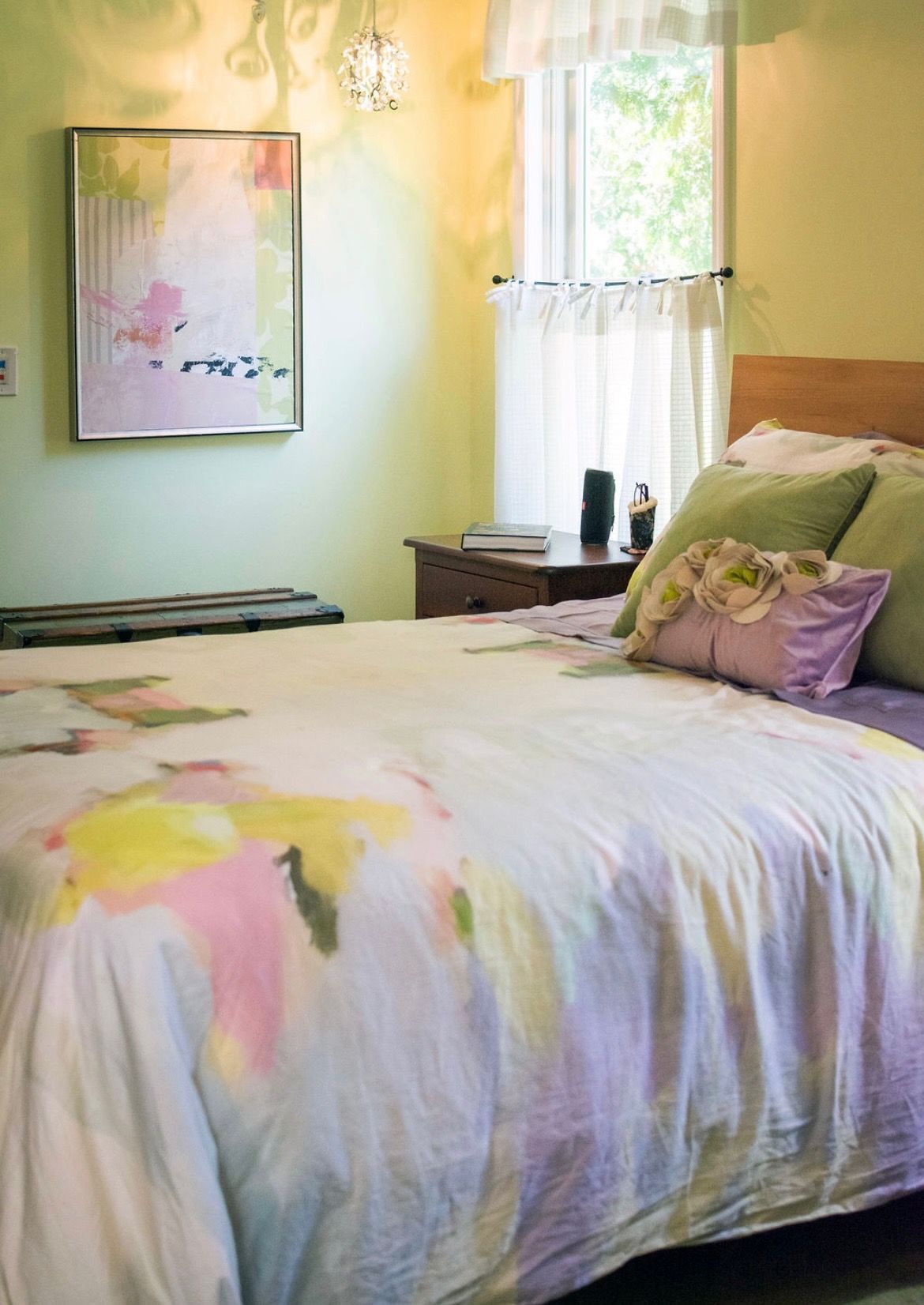 A bedroom with a pale green wall, a bed covered in a watercolor-patterned quilt, and a framed abstract painting.