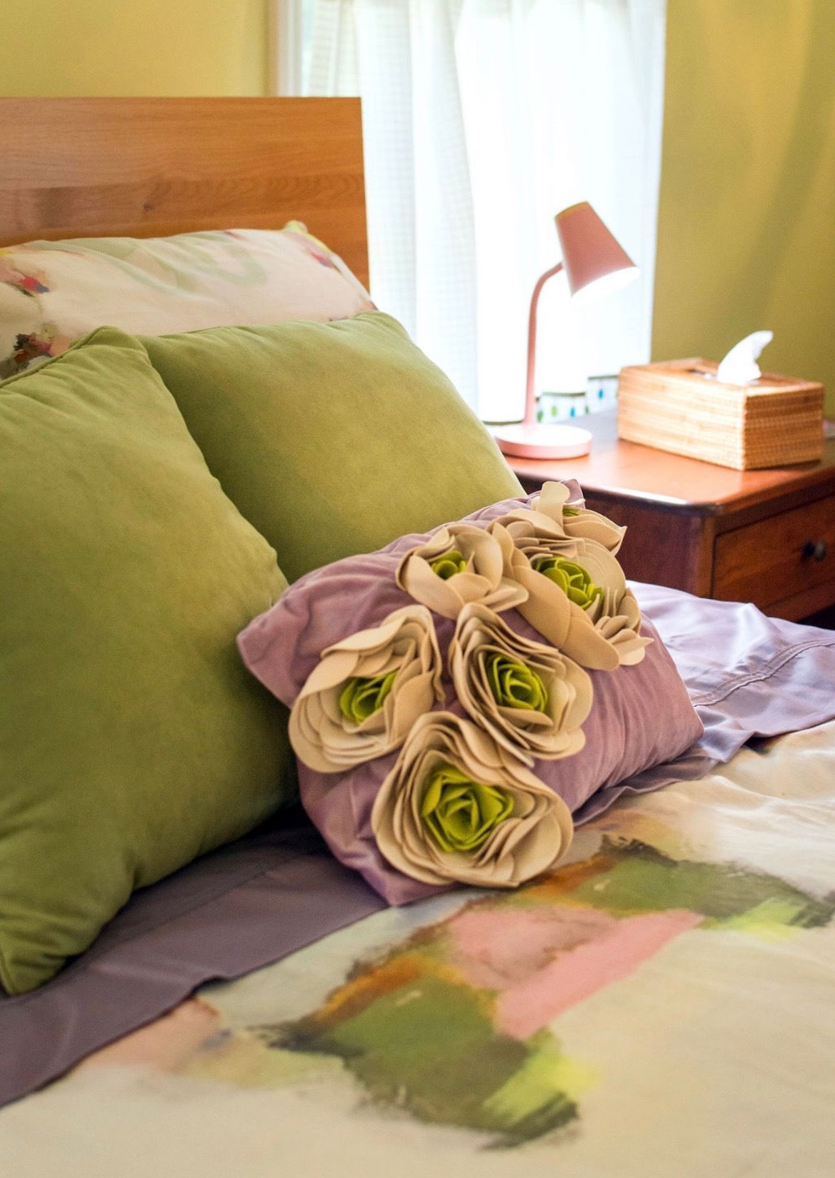 A cozy bed with green pillows, a purple throw pillow with fabric flowers, and a bedside lamp with a wooden tissue box.