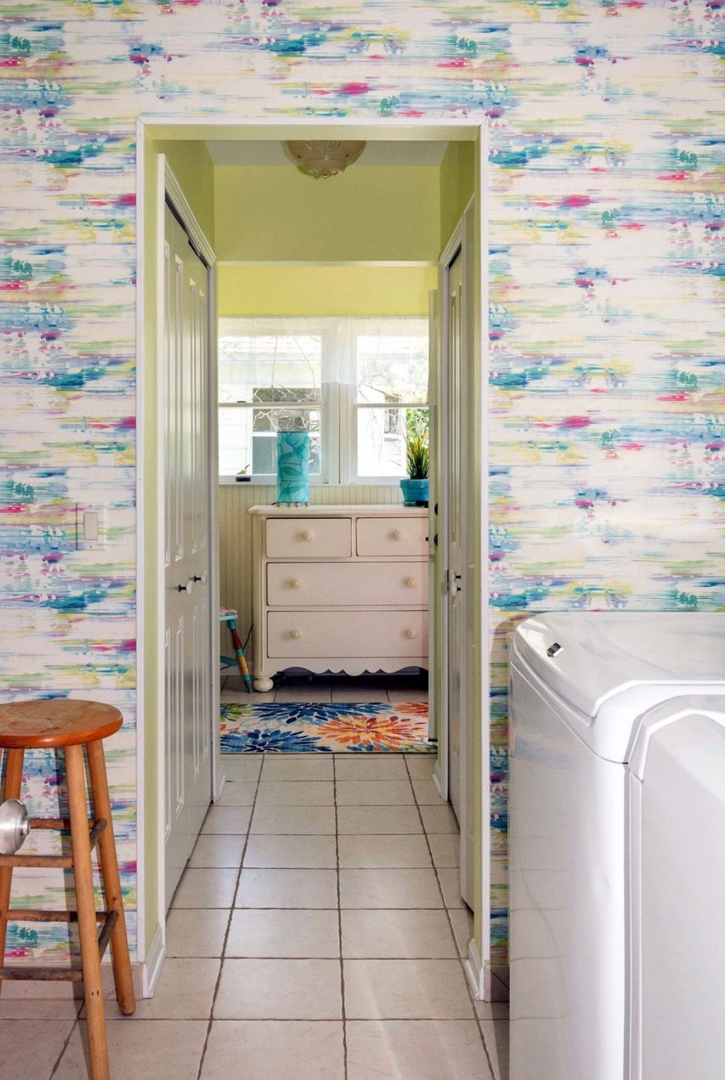 A laundry room with patterned wallpaper, tile floor, wooden stool, and a doorway leading to a sunlit room with a dresser.