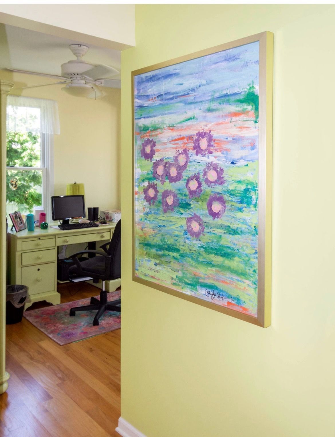 A framed abstract floral painting hangs on a light green wall, with a home office visible through a doorway beyond.