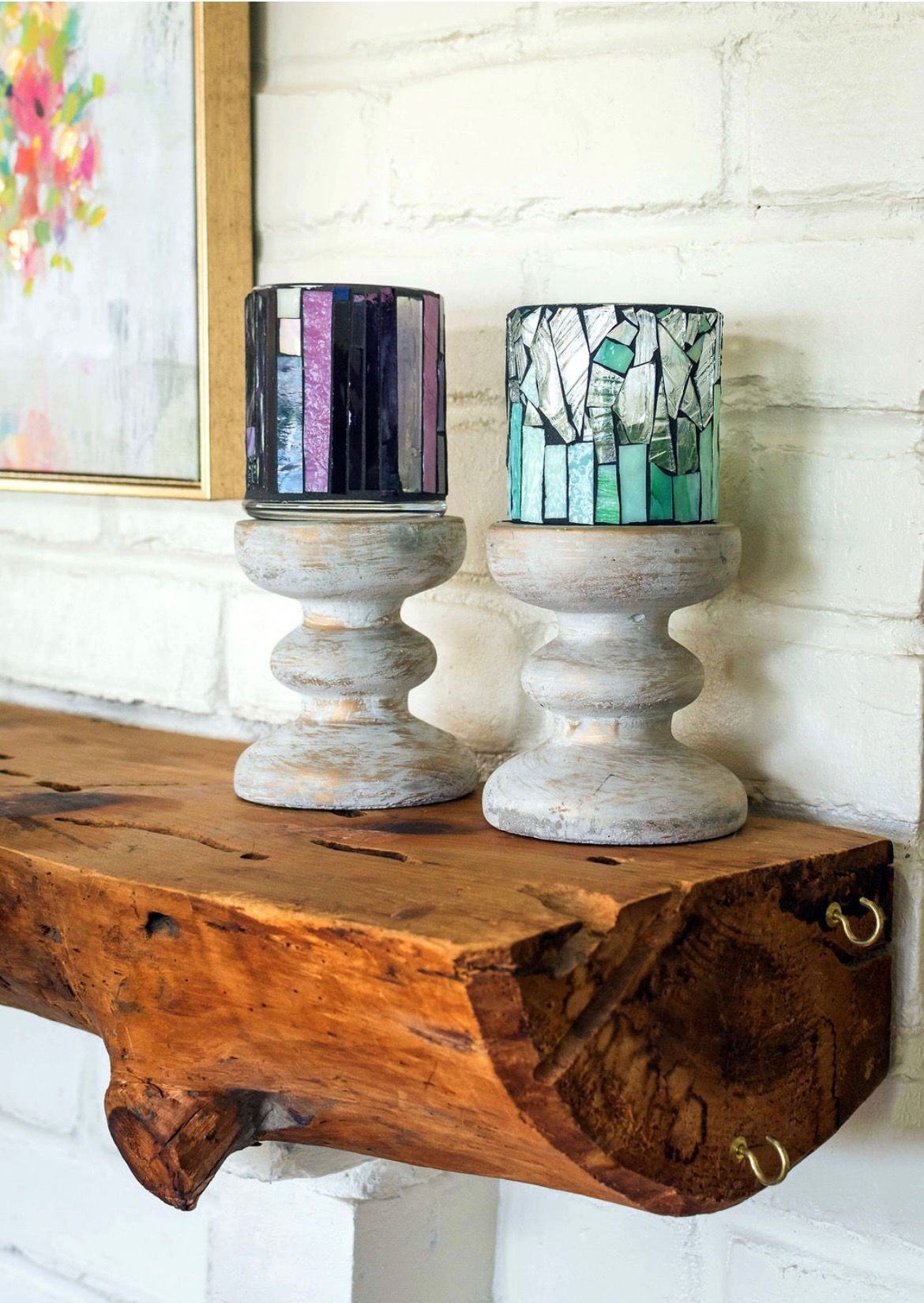 Two decorative glass candle holders on rustic wooden stands sit on a log mantle against a white brick wall.