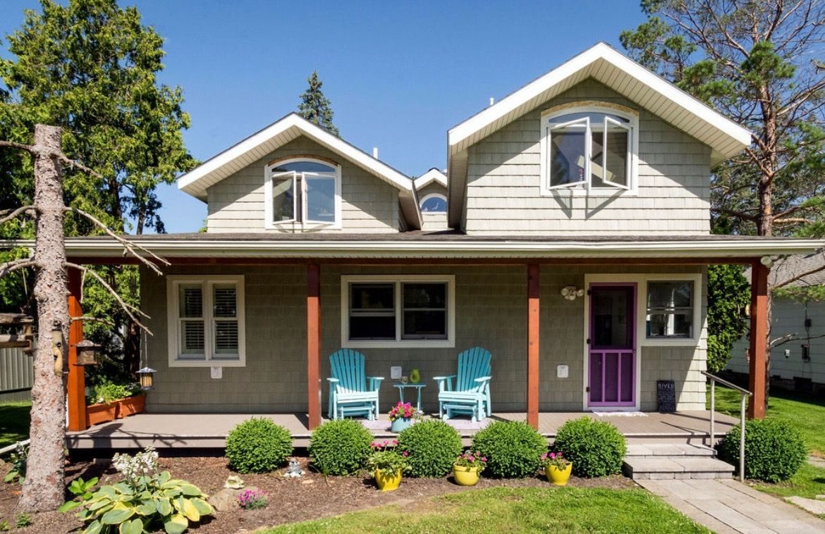 A two-story green house with a front porch, featuring two blue Adirondack chairs, a purple door, and manicured shrubs.