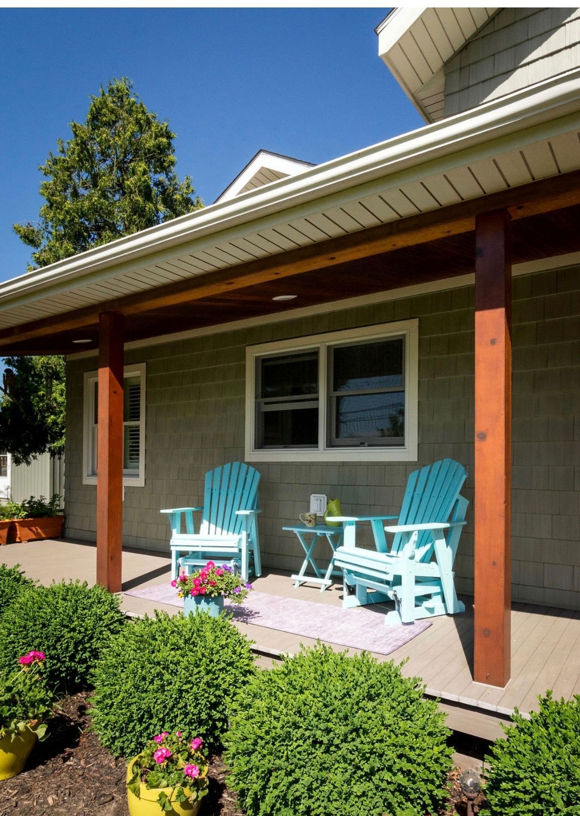 Two turquoise Adirondack chairs and a small table sit on a porch of a home with green shingles and wooden pillars.