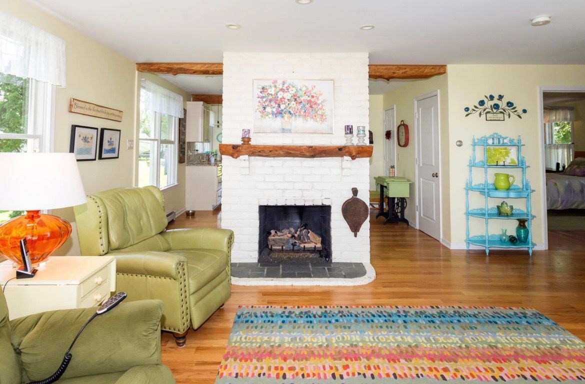 A living room with a white brick fireplace, green armchair, colorful rug, and light wood floors.