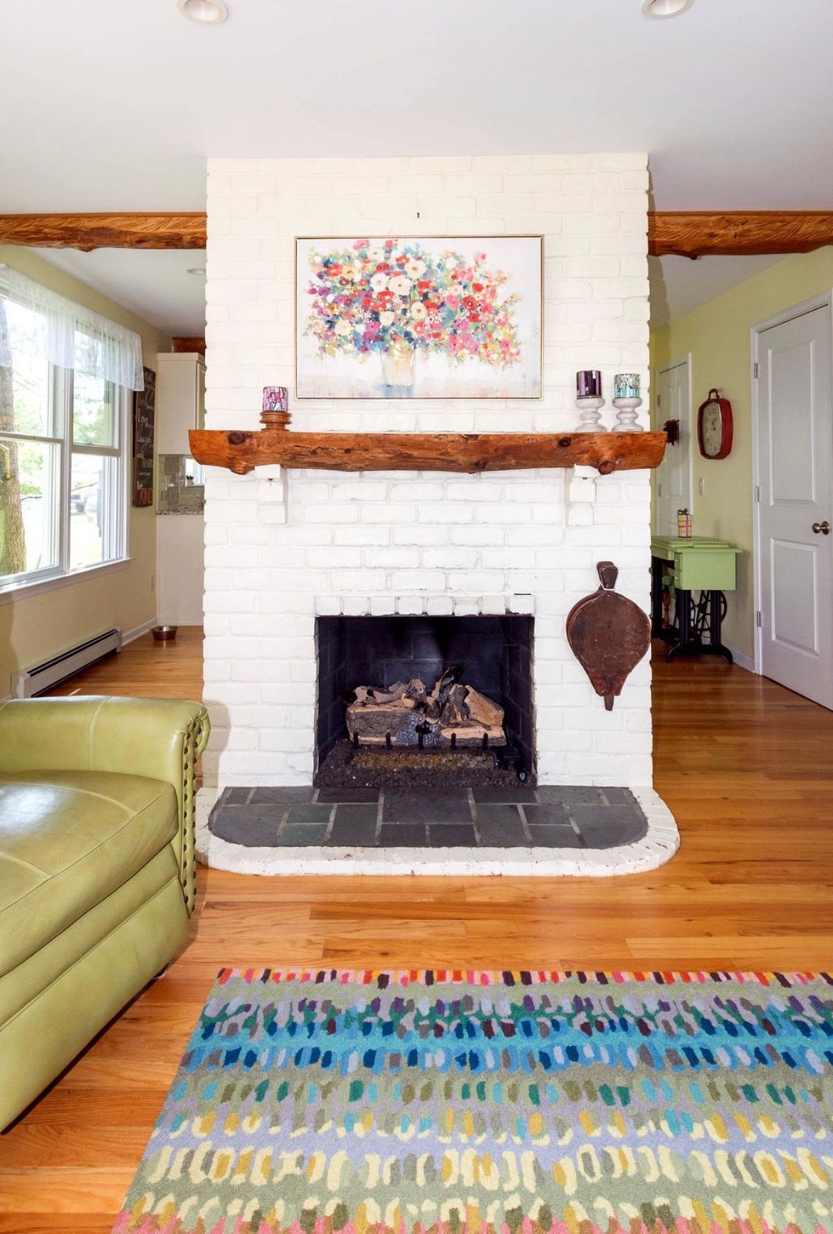 A white painted brick fireplace with a rustic wood mantel, colorful abstract artwork, and a patterned rug in front.