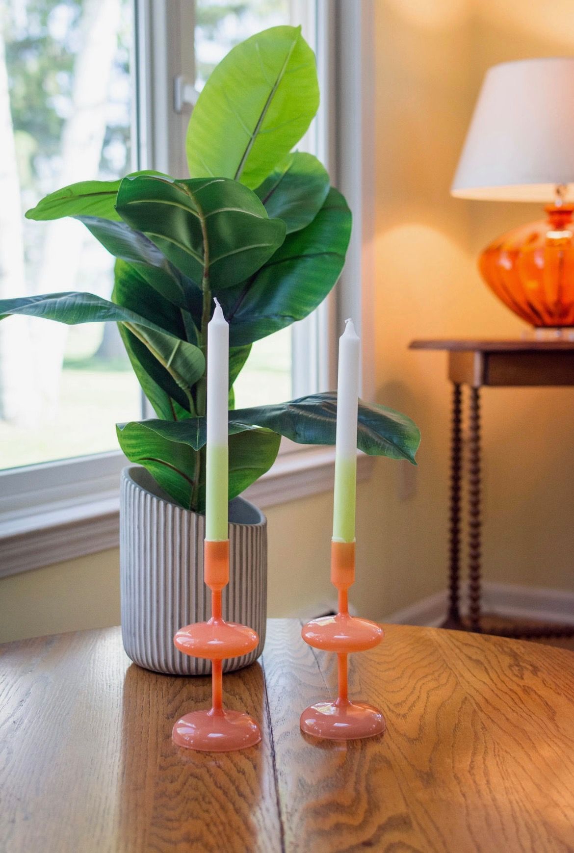Two ombre orange glass candlesticks with white candles sit on a wooden table beside a leafy potted plant.