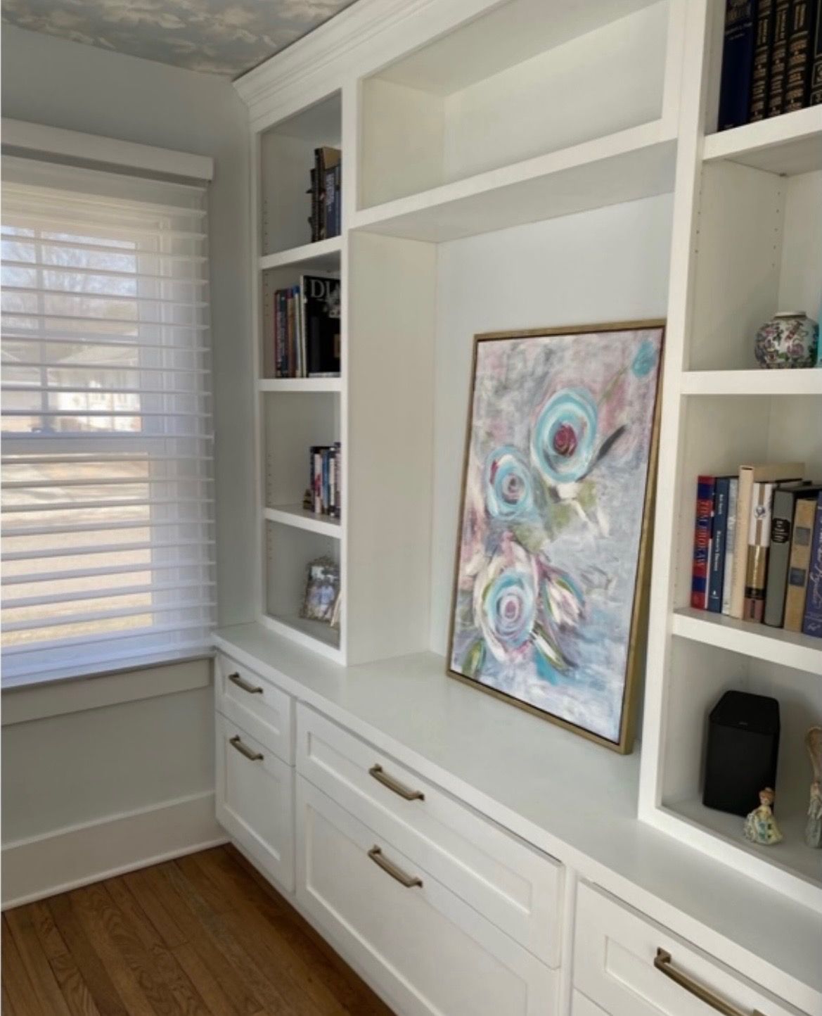 White custom built-in shelves and cabinets next to a window with white blinds, featuring an abstract floral painting.