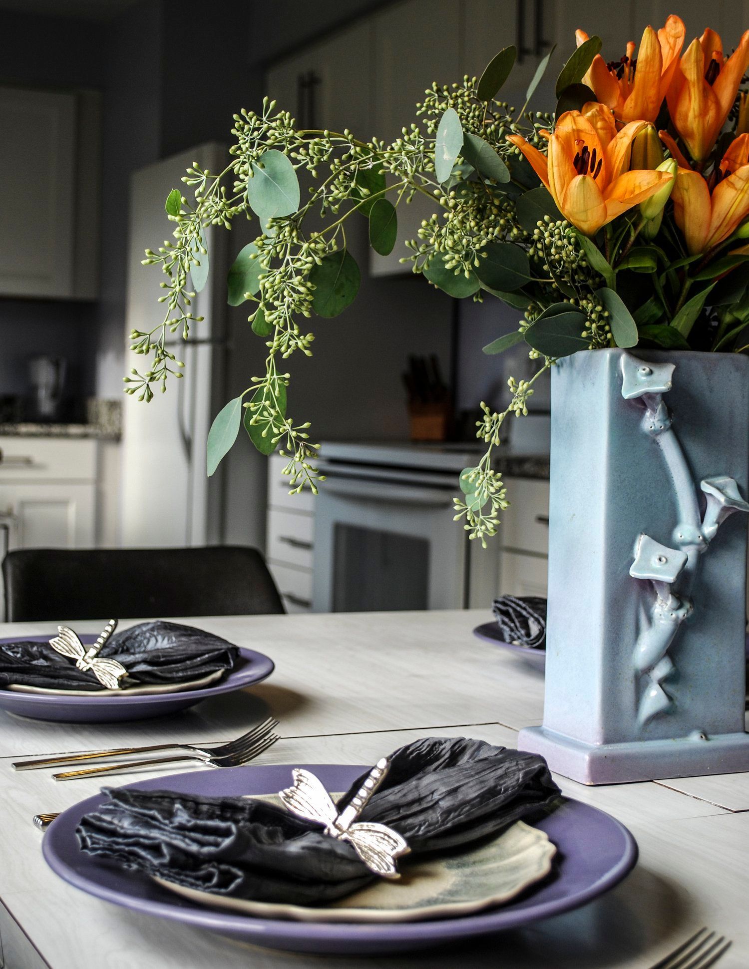 A table setting with purple plates, dark napkins with dragonfly rings, and a vase of orange lilies in a kitchen.