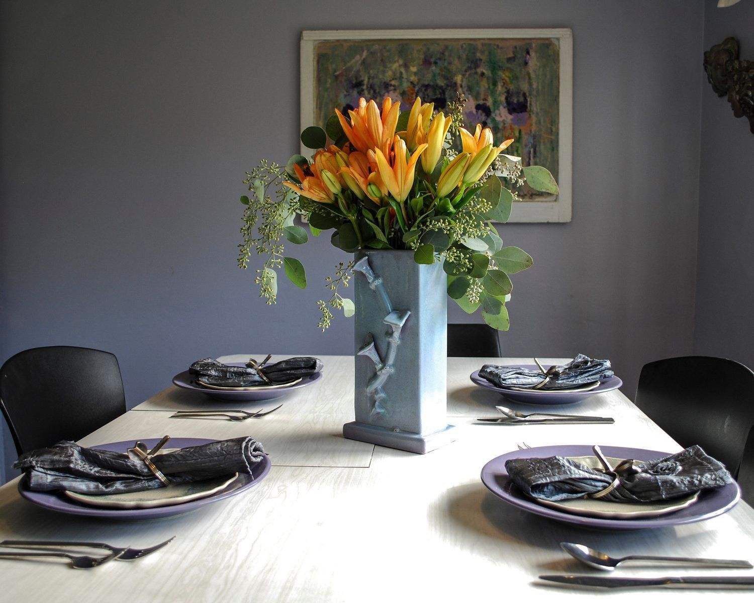 A dining table set for four with light-purple plates, dark gray napkins, and a blue vase with yellow lilies as a centerpiece.